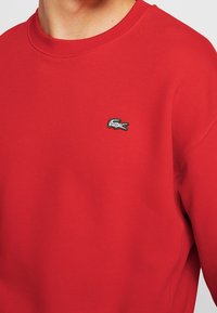 Primo piano di una persona che indossa un felpa rossa con girocollo, decorata con un piccolo logo di coccodrillo verde e bianco sul petto.