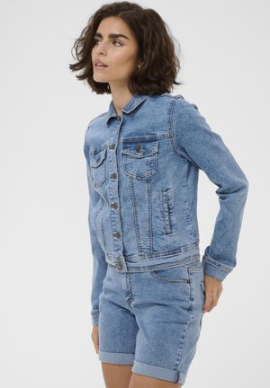 Femme aux cheveux courts et foncés portant une veste en jean bleu clair et un short assorti retroussé, debout devant un fond clair uni.