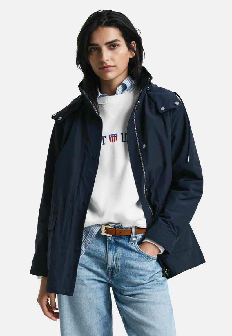 Marinblå vattentät jacka med huva, frontdragkedja och tryckknappar, lager på en vit sweatshirt och blå jeans. Har en slät yta.