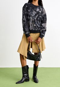 Musta batikkipainettu collegepaita, jossa on resorit hihansuissa, khakinvärinen epäsymmetrinen hame, musta nahkainen käsilaukku ja polven korkeudelle ulottuvat mustat saappaat.