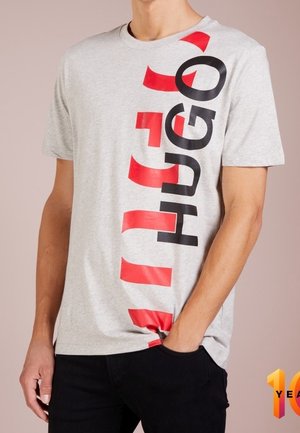 Mand iført lysegrå T-shirt med korte ærmer og stort vertikalt "HUGO"-logo i sort og røde bogstaver, kombineret med sorte bukser.