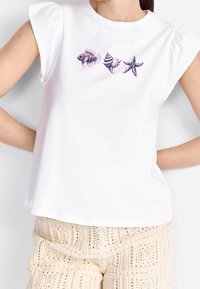Camiseta blanca de manga corta con mangas abullonadas, que presenta gráficos de conchas moradas y estrellas de mar en la parte delantera. La tela parece suave y lisa.