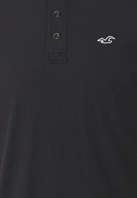 Melna polo krekls, izgatavots no kokvilnas, ar pogu slēgšanu un izšūtu logo. Vienkāršs dizains, mīksta tekstūra, pie apkakles ir trīs pogas.