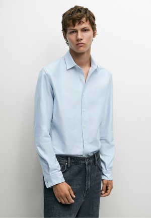 Giovane uomo con capelli castani corti e ricci che indossa una camicia a righe azzurro chiaro con bottoni e jeans scuri, in piedi davanti a uno sfondo bianco semplice.