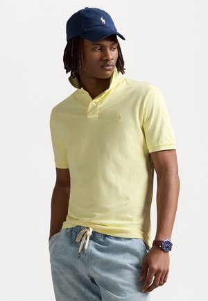 Gul polo shirt med kravehalsudskæring, korte ærmer og broderet logo, parret med lyseblå shorts og en mørkeblå kasket.