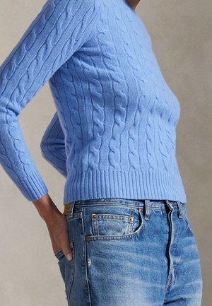 Personne portant un pull torsadé bleu clair et un jean bleu, avec une main dans la poche arrière.