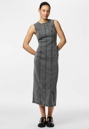 Noor naine halli, varrukateta, keha järgi istuvat midikleit koos vertikaalsete õmblustega, seisab käed selja taga, seljas mustad pandlaga kingad.