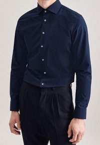 Camicia a maniche lunghe blu navy in tessuto liscio, con chiusura a bottoni, colletto classico e una leggera lucentezza. Abbinata a pantaloni neri plissettati.