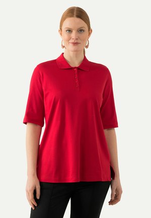 Camisa polo roja de manga corta, con cuello y una fila de botones. Tejido suave con un corte relajado y aberturas laterales. Llevada con pantalones negros.