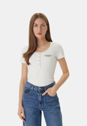 Donna con lunghi capelli castani che indossa una maglietta henley bianca a maniche corte e jeans blu, in piedi con una mano in tasca, espressione neutra.