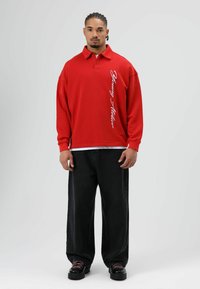 Rode polo met lange mouwen en een kraag; heeft witte verticale tekst. Gecombineerd met wijde zwarte jeans en zwarte schoenen met roze veters.