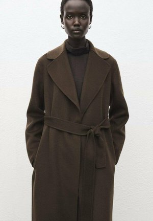 Cappotto lungo marrone con rever a punta, cintura intrecciata e texture liscia. Indossato sopra una maglietta nera, caratterizzata da un design minimalista.