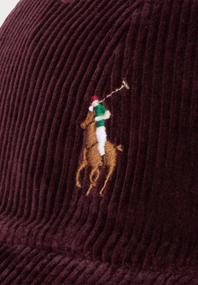Burgunderrød kordfløjelscaps med et broderet polo-spillerlogo i guld, grøn og rød. Visuelt struktureret med tydelige ribber.
