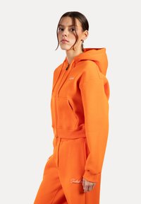 Haut cropped orange vif avec fermeture éclair à l'avant, poignets côtelés et deux poches avant. Présente un logo de marque blanc sur la poitrine.