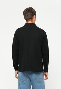 Black long-sleeve shirt with a collar, featuring button details on the cuffs, and a clean, smooth fabric texture. Worn with light blue jeans.