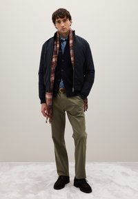 Giovane uomo in piedi con le mani in tasca, indossa una giacca blu navy, una camicia blu, un cardigan nero, pantaloni oliva, una sciarpa a scacchi e scarpe scure.