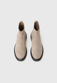 Bottines en daim beige avec une semelle en caoutchouc noir, présentant un design minimaliste et une languette à l'arrière pour un enfilage facile.