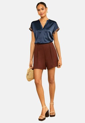 Kvinde iført en marineblå satinbluse med leopardkanter, brune shorts med høj talje, beige platåsko og holder en lille vævet håndtaske.