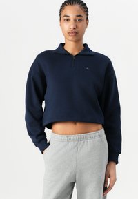 Marineblå cropped genser med glidelås og høy krage, laget av mykt stoff. Kombinert med lysegrå joggebukser i avslappet passform.