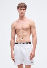 Λευκά μαγιό σορτς με μαύρη ελαστική μέση με λογότυπο, κορδόνι και άνετη γραμμή, φορεμένα από μοντέλο με γυμνό κορμό.