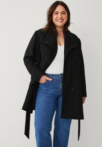 Sort uldblandings trenchcoat med bred krave, sidelommer og bindebånd i taljen. Båret sammen med en hvid top og blå jeans.