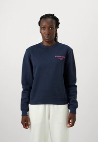 Tengerészkék pulóver puha anyagból, kerek nyakkivágással, bordázott mandzsettákkal és rózsaszín logóval a bal mellrészen.