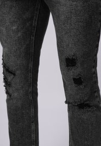Jeans de mezclilla gris oscuro con áreas desgastadas, bordes deshilachados y grandes desgarrones. Costuras visibles a lo largo de los lados. Diseño ajustado.