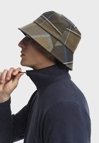 Cappello a secchiello in tessuto a quadri marrone, grigio e blu. Giacca con colletto in pile. Il soggetto chiude la giacca con una mano.
