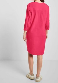 Robe rose, arrivant au genou, fabriquée à partir d'un tissu texturé avec des manches trois-quarts. Elle présente un col arrondi et des poignets boutonnés. Associée à des baskets beiges.