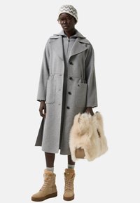 Kvinde iført grå frakke, strikket hat, briller og beige pelsforede støvler, der holder en beige fluffy håndtaske mod en hvid baggrund.