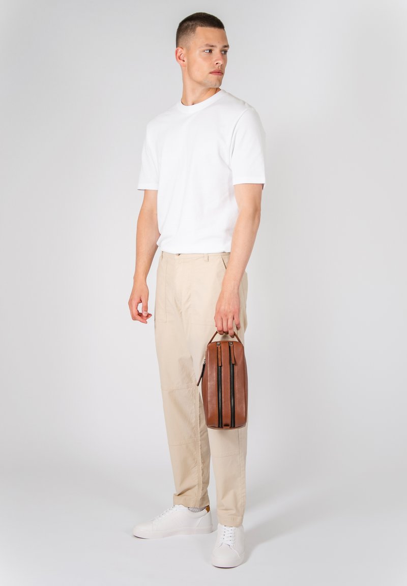Borsa in pelle marrone con doppia cerniera, indossata da una persona in t-shirt bianca e pantaloni beige, in piedi su uno sfondo neutro.