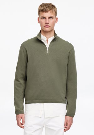 Šviesiaplaukis vyras, dėvintis alyvuogių žalią ketvirčio užtrauktuko megztinį ant baltos marškinių su sagomis ir baltų kelnių, stovintis prie balto fono.