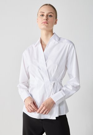 Weiße Langarm-Wickelbluse mit spitzem Kragen, Kordelzug in der Taille und glattem Baumwollstoff; kombiniert mit schwarzen Hosen.