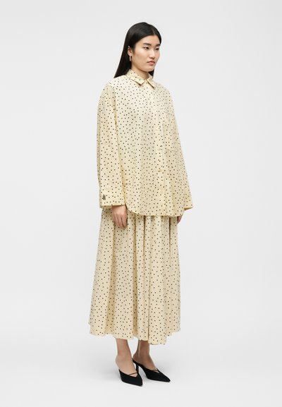 Femme debout portant une chemise beige à manches longues à pois et une jupe longue assortie, associées à des chaussures à talons noires.