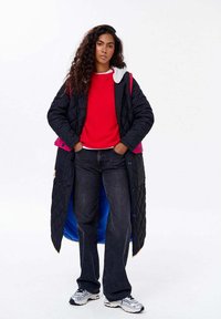 Žena s dlouhými kudrnatými vlasy, oblečená v černém prošívaném kabátu s modrým podšívkou, červeném svetru, černých džínách a bílých teniskách, stojí se založenýma rukama v kapsách.