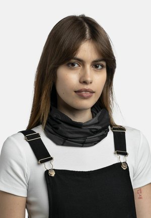 Young woman with straight brown hair wears white short-sleeve shirt, black overalls, and a black neck gaiter, facing forward with neutral expression.