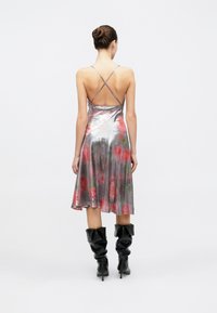 Robe nuisette métallique avec un ourlet évasé, présentant un motif floral aux tons rouges et verts. Bretelles croisées ajustables au dos. Bottes noires montant jusqu'aux genoux.