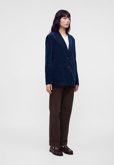 Femme portant un blazer bleu marine, un haut blanc, un pantalon marron et des mocassins marron foncé, debout sur un fond clair uni.