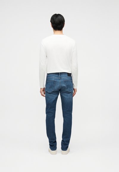 Henkilö seisoo selin päin, päällään valkoinen pitkähihainen paita, siniset farkut takataskuilla ja vaaleat lenkkarit yksivärisellä taustalla.