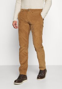 Bruine corduroy broek met verticale ribbels, smalle pasvorm en voorzakken. Gecombineerd met donkergroene leren laarzen. Textuuropppervlak zichtbaar.