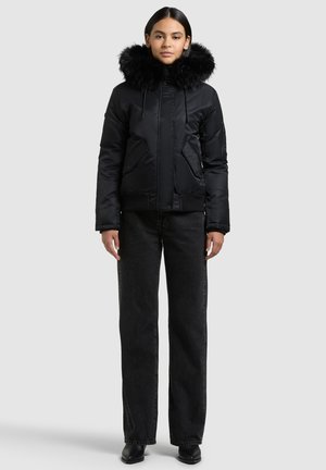 Giacca a vento nera imbottita con cappuccio in pelliccia sintetica staccabile, chiusura con zip e tasche frontali, indossata con jeans neri a vita alta e stivali neri.