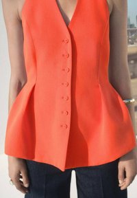 Red peplum top in textured fabric with a deep V neckline, featuring six front buttons and flared sides, paired with dark denim pants.