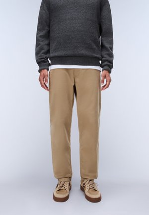 Personne portant un pantalon beige, des baskets beige et marron, une chemise blanche et un pull gris foncé, se tenant devant un fond uni.
