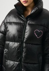 Mujer vistiendo una chaqueta de plumas negra brillante con un corazón rosa y el texto "Laurella" en el lado izquierdo del pecho, con la cremallera subida.