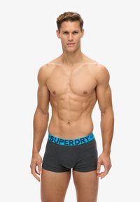 Pantalones cortos grises con una cinturilla elástica azul que presenta la marca "SUPERDRY". La tela parece suave, ofreciendo un ajuste ceñido.