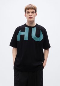 Μαύρο βαμβακερό μπλουζάκι με μεγάλο τουρκουάζ γραφικό «HU» στο μπροστινό μέρος, σε άνετη γραμμή, με στρογγυλή λαιμόκοψη και κοντά μανίκια.