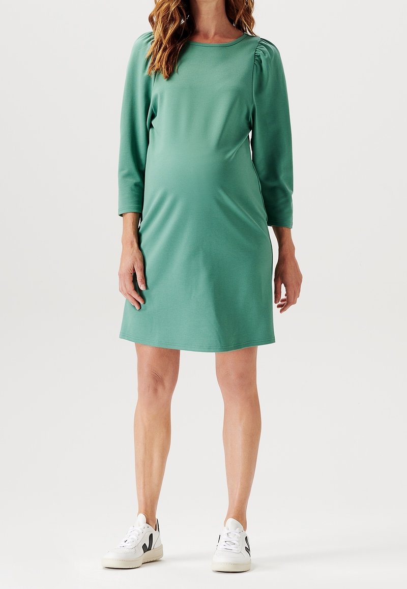 Femme enceinte portant une robe verte mi-longue avec des manches trois-quarts et des baskets blanches, debout sur un fond uni.