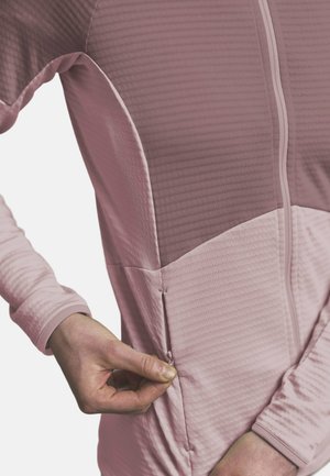 Person wearing a two-tone pink zip-up jacket, pulling the small zipper on the side pocket with their right hand.