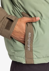 Grön isolerad jacka med vertikalt ribbat mönster, med khaki-accenter och en Quiksilver-logotyp på sidofickan. Kardborrefäste på ärmen.