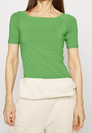 Mujer que lleva un top verde ajustado de manga corta con escote amplio y pantalones crema de talle alto con una gran cintura doblada.